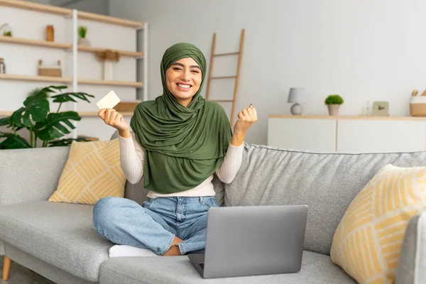 Joven dama árabe alegre en hijab gesto SI, la celebración de la tarjeta de crédito, compras en la tienda en línea en el ordenador portátil en casa — Foto de Stock