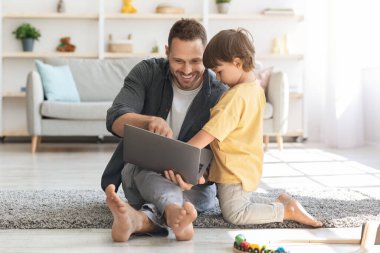 Modern çocukların ilgi alanları. Sevimli küçük çocuk babasının dizüstü bilgisayarda çalışmasını izliyor, neşeli bir adam oğluna nasıl çevrimiçi çalışacağını gösteriyor.