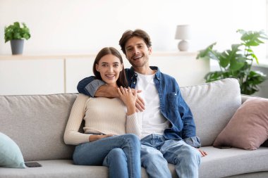 Neşeli bin yıllık beyaz bir adam, bir kadına kanepede sarılıyor ve oturma odasında kameraya bakıyor.