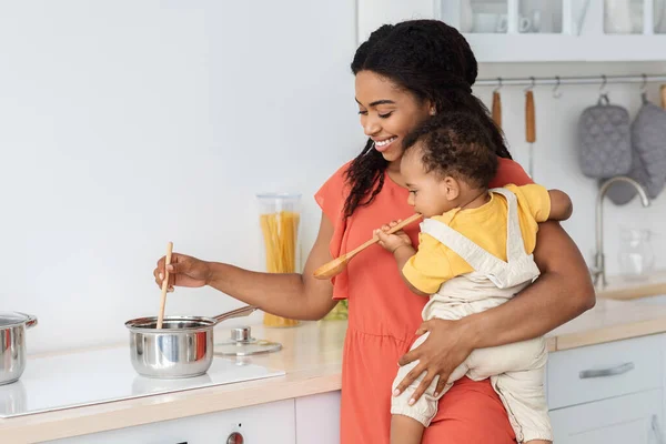 Glückliche junge schwarze Mutter kocht in der Küche mit kleinem Baby auf Händen — Stockfoto