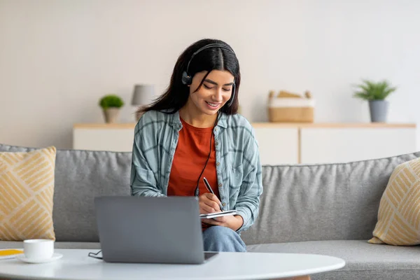 Jonge Arabische vrouw in hoofdtelefoon met behulp van laptop, werken of studeren online van huis, notities maken tijdens zakelijke bijeenkomst — Stockfoto