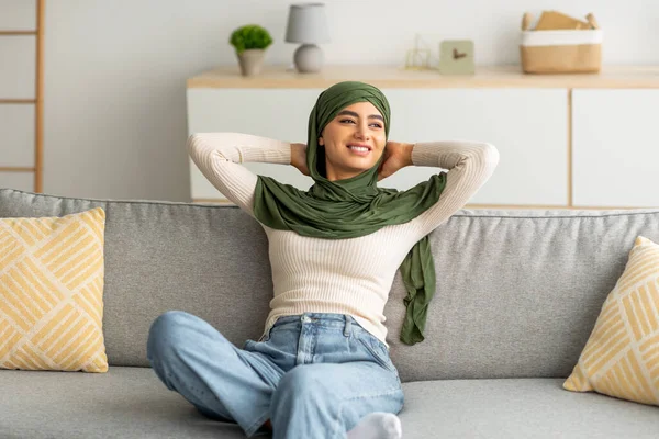 Wanita Arab muda yang cantik mengenakan jilbab duduk di sofa dengan tangan di belakang kepala, beristirahat dan bersantai di rumah — Stok Foto