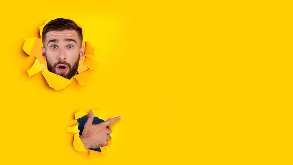 Sorprendido joven caucásico con barba mira a través del agujero en papel amarillo y señala el dedo para liberar espacio — Foto de Stock