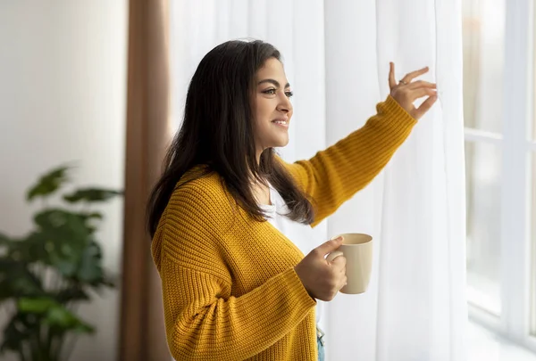 若い中東の女性が窓のそばに立ってお茶のマグカップ、カーテンを移動し、窓を通して見て笑顔 — ストック写真
