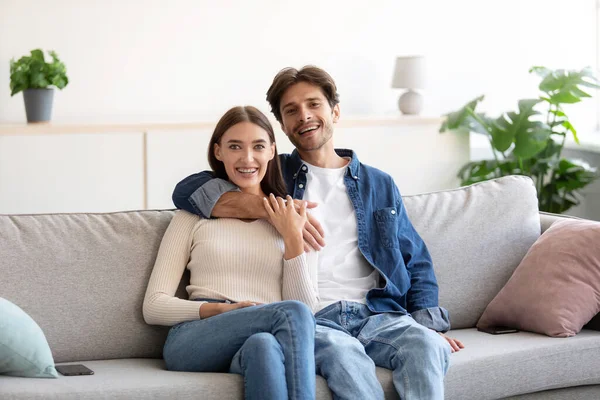 Feliz alegre milenial caucásico chico abraza a la mujer en el sofá y mirando a la cámara en la sala de estar —  Fotos de Stock