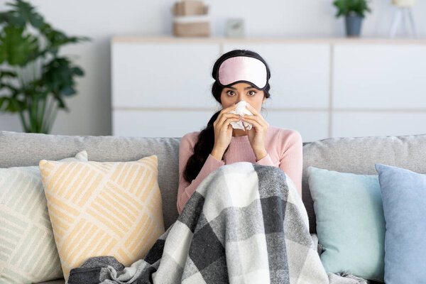 Young pretty hindu woman in sleep mask covered in blanket, suffers from flu, blows nose in napkin