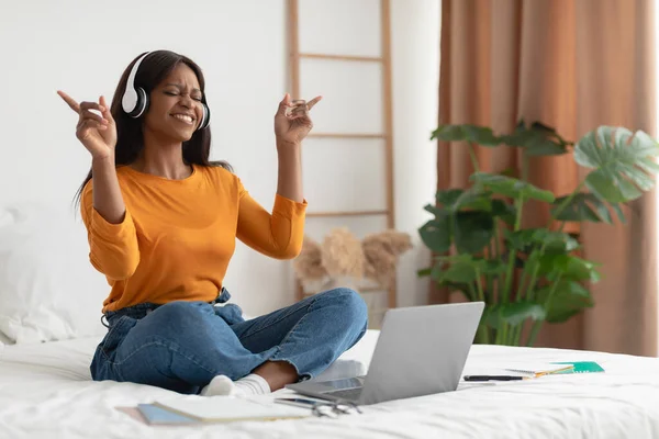 Yatak odasında dizüstü bilgisayar ve kulaklık kullanarak müzik dinleyen siyah kadın. — Stok fotoğraf