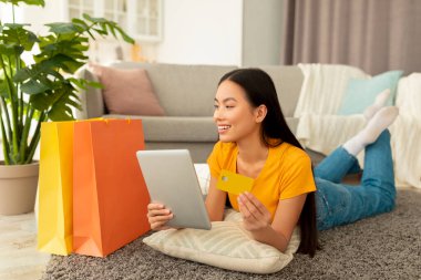 Alışverişte ve e-ticarette. Kredi kartı tutan pozitif Asyalı kadın, tabletle alışveriş yapıyor, yerde yatıyor.