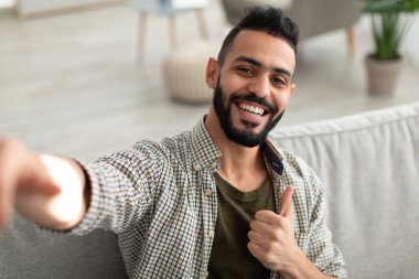 Mutlu genç Arap adam selfie çekiyor ve parmağını kaldırıyor, cep telefonu kamerasına gülümsüyor, evdeki kanepede oturuyor.