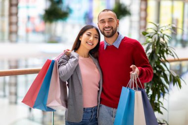Alışveriş torbalarıyla poz veren, büyük alışveriş merkezinde kameraya gülümseyen mutlu çiftin portresi.