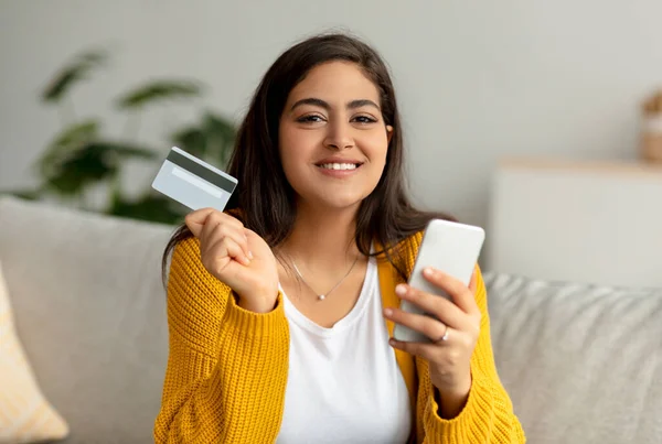 Çevrimiçi ödeme kavramı. Elinde kredi kartı ve akıllı telefon tutan gülümseyen müslüman kadın, koltukta oturuyor ve kameraya bakıyor. — Stok fotoğraf