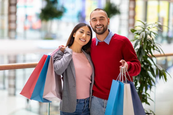 Portret van jong gelukkig gevarieerd koppel poseren met shopper tassen, glimlachen op camera in enorme stad winkelcentrum — Stockfoto
