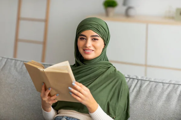 Evdeki hobilerin rahat olsun. Türbanlı genç Arap kadın kitap okuyor, oturma odasında kanepede oturuyor. — Stok fotoğraf