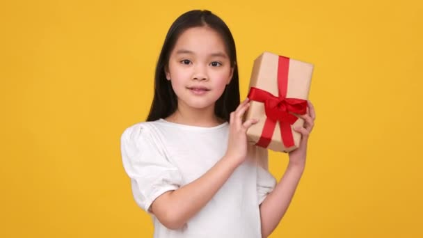 Semester nyfikenhet. bedårande liten asiatisk flicka skakar presentförpackning, försöker gissa vad inuti, orange studio bakgrund — Stockvideo
