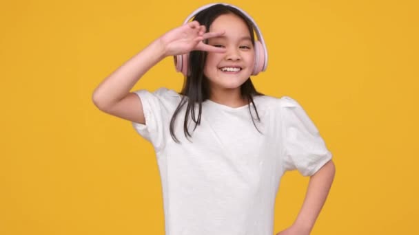 Glückliche Stimmung. Fröhliches hübsches asiatisches kleines Mädchen hört Musik in großen rosafarbenen drahtlosen Kopfhörern und tanzt vor der Kamera — Stockvideo