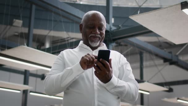 Зрелый черный бизнесмен с помощью смартфона, стоя в современном офисном центре в помещении — стоковое видео