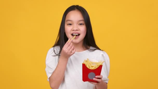 Aperitivos favoritos para niños. Linda chica asiática comiendo papas fritas del paquete de comida para llevar, disfrutando de la comida rápida sabrosa chatarra — Vídeos de Stock