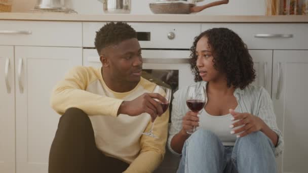 Jóvenes cónyuges afroamericanos positivos discutiendo noticias, tintineo con copas de vino tinto, relajarse en el suelo de la cocina — Vídeos de Stock