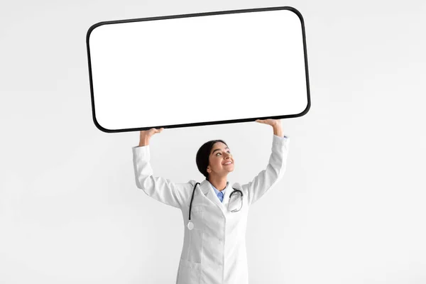 Feliz milenial mujer india médico en blanco abrigo celebrar teléfono grande con pantalla en blanco por encima de la cabeza —  Fotos de Stock