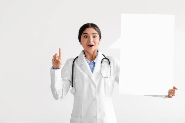 Sorprendido alegre milenial mujer india médico en abrigo sostiene espacio en blanco abstracto para las palabras y muestra el dedo hacia arriba —  Fotos de Stock