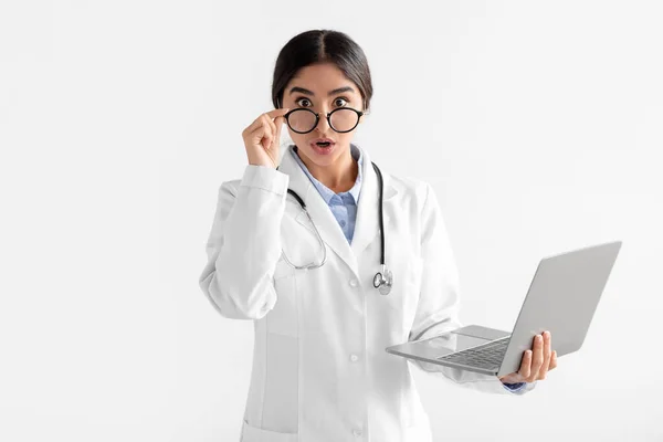 Sorpreso giovane medico indù femminile in camice bianco toglie occhiali e guarda la macchina fotografica, lavora al computer portatile — Foto Stock