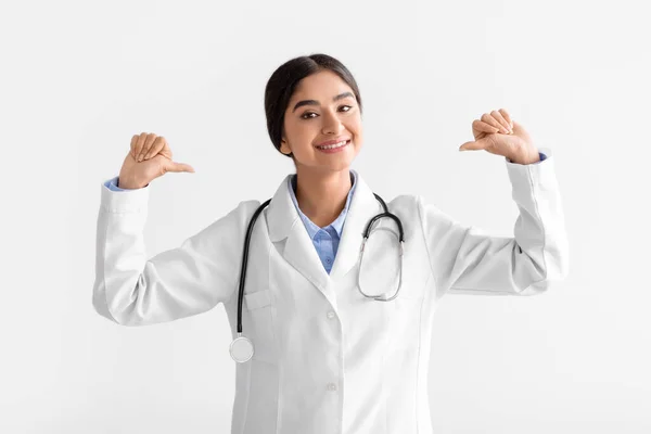 Alegre sonriente joven india doctora en abrigo señala los dedos a sí misma, aislada sobre fondo blanco, estudio —  Fotos de Stock