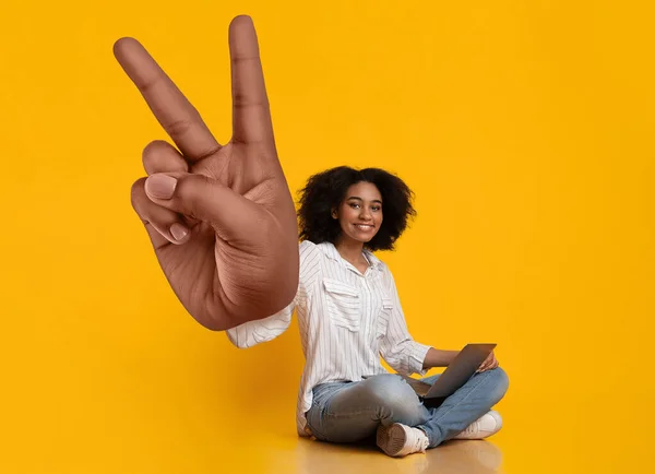 Cool young black lady sitting with laptop computer, showing big peace or victory gesture on orange studio background — Stock Photo, Image