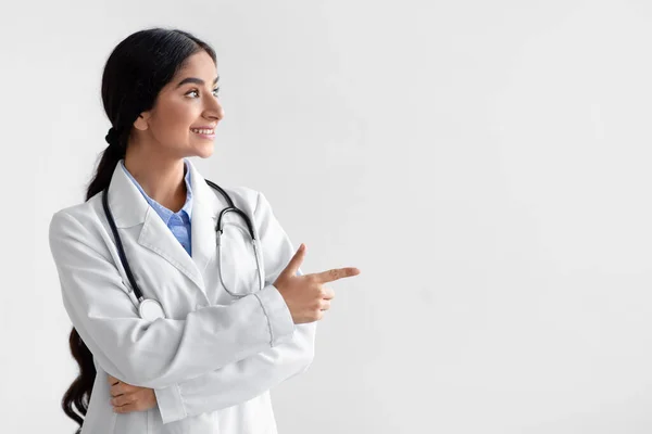 Smiling millennial indian woman doctor, family therapist in white coat show finger an empty space — Stock Photo, Image