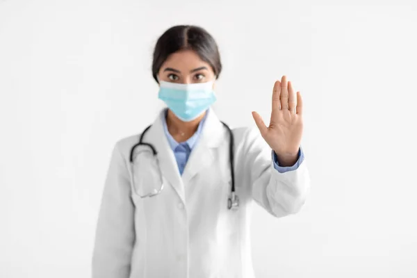 코트를 입고 보호 마스크를 쓰고 있는 진지 한 젊은 인도 여성 의사가 스톱 핸드 동작을 보여 줍니다. — 스톡 사진