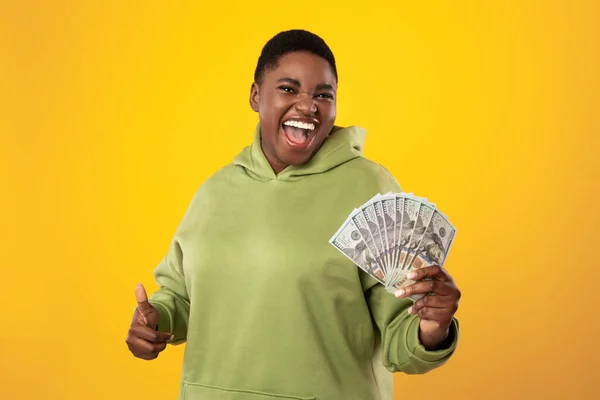 Счастливая афроамериканка держит в руках деньги, держа пальцы вверх, на желтом фоне — стоковое фото