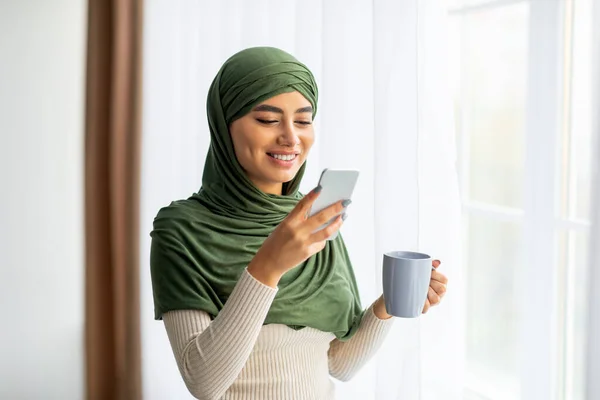 Happy young Arab woman in hijab standing near window with coffee and smartphone, browsing internet, chatting online — Stock Photo, Image