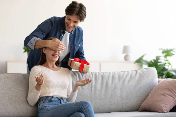 Sourire millénial européen gars ferme les yeux à la femme, donne présent dans la boîte pour surprendre femme souriante — Photo