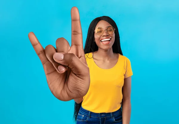 Mulher afro-americana alegre mostrando grande gesto de rock, sorrindo para a câmera, usando óculos de sol no fundo azul — Fotografia de Stock