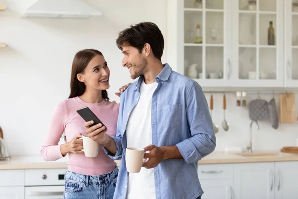 Mosolygó boldog millenniumi női és férfi csésze forró ital beszélgetés a modern konyhában — Stock Fotó