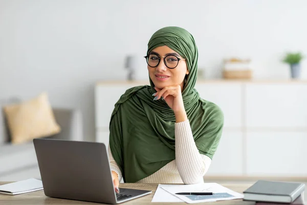 Genç Arap kadının tesettürlü portresi dizüstü bilgisayar kullanıyor, masasında oturuyor, internette çalışıyor, evde giysi giyiyor. — Stok fotoğraf