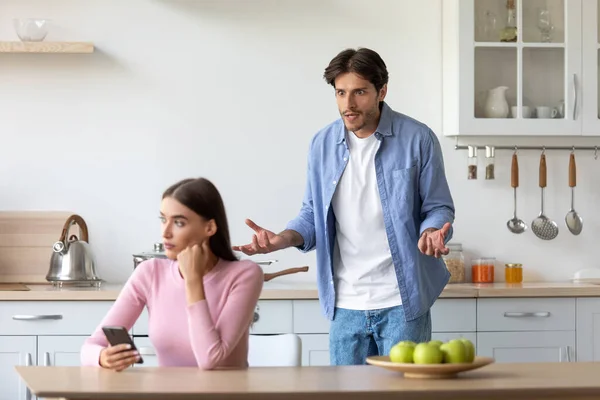 Sad angry young man yelling at woman with smartphone, unhappy lady ignoring him on minimal kitchen