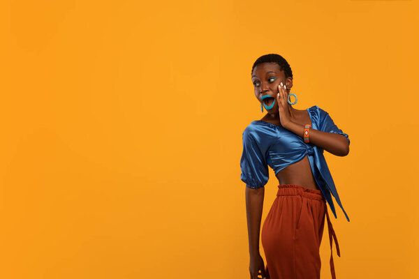 Amazed attractive african american millennial woman in bright stylish outfit and makeup looking at copy space with open mouth and touching her face over yellow studio background