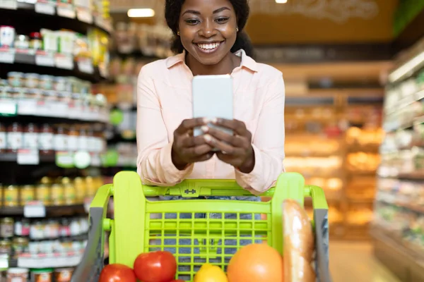 スーパーで食料品の買い物中に携帯電話を使用して幸せな黒の女性 — ストック写真