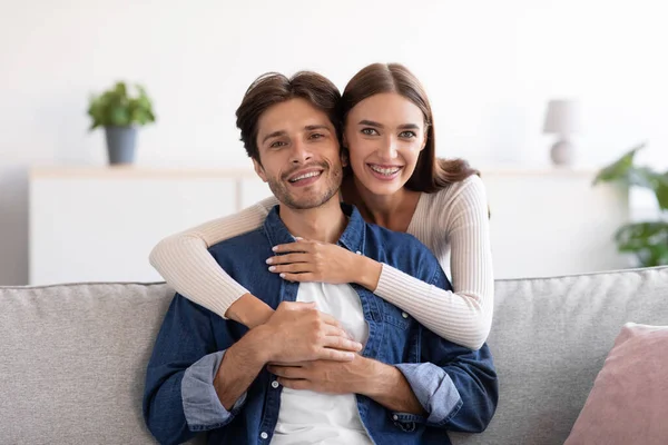 Feliz jovem esposa abraços marido por trás no sofá e olhar para a câmera no interior do quarto — Fotografia de Stock