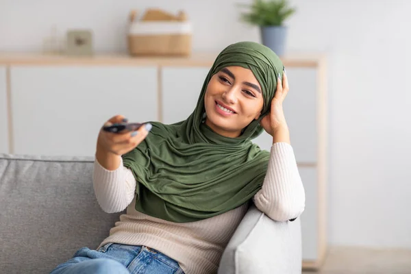 穆斯林头巾中的年轻阿拉伯妇女手持遥控器，在家里沙发上看电视 — 图库照片