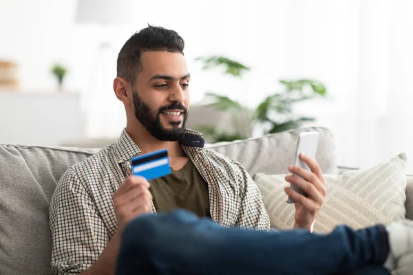 Vonzó fiatal arab férfi mobiltelefonnal és hitelkártyával online vásárláshoz vagy távoli banki ügyintézéshez otthon — Stock Fotó