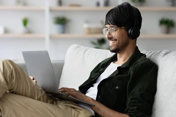 Positivo ragazzo indiano che si rilassa a casa, utilizzando laptop e auricolari — Foto Stock