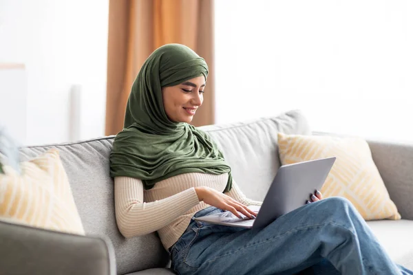 Портрет молодой арабской женщины в хиджабе, сидящей на диване, использующей ноутбук, серфинг в Интернете, работающей или учащейся дистанционно дома — стоковое фото