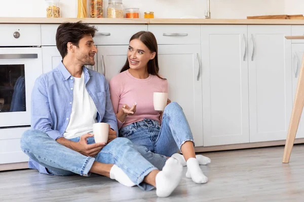 Feliz milenar caucasiano feminino e masculino com xícaras de bebida, sentado no chão, falando na cozinha — Fotografia de Stock