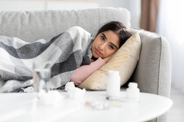 Frustrace mladá hinduistka pokrytá kostkovanou, trpí chřipkou, leží na pohovce v interiéru pokoje s vodou a pilulky — Stock fotografie