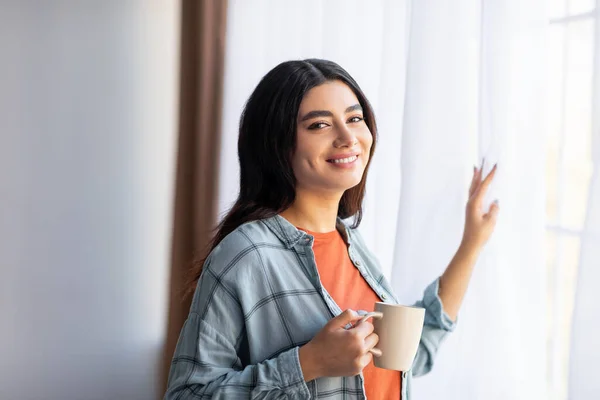 Gelukkig jong Arabisch dame drinken van verse koffie in de buurt venster thuis, genieten van ontspannen ochtend — Stockfoto