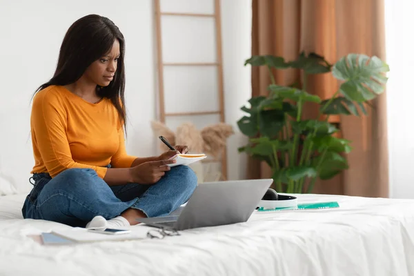 Umtriebige schwarze Frau macht sich Notizen beim Lernen mit Laptop im Schlafzimmer — Stockfoto