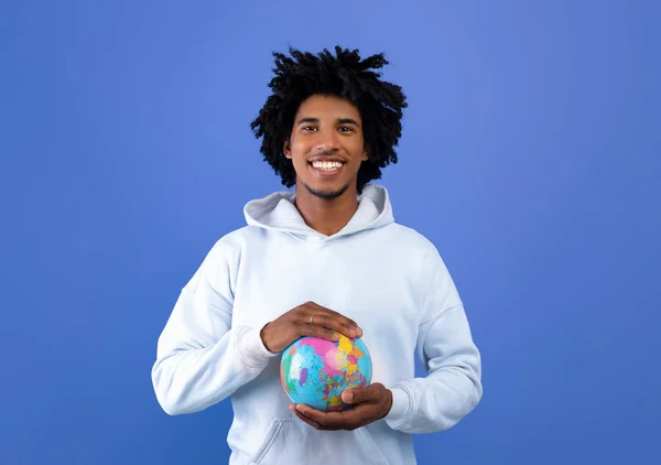 Feliz adolescente negro protegendo globo com as mãos no fundo do estúdio azul. Ecologia, turismo, igualdade da humanidade — Fotografia de Stock