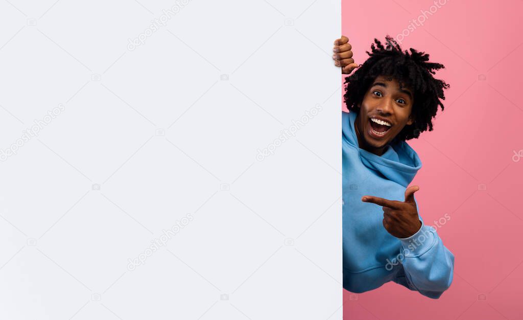 Excited African American teenager hiding behind empty paper poster with mockup for your ad design on pink background