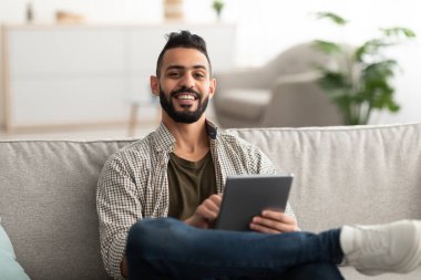 Evdeki kanepede otururken dijital tablet kullanan yakışıklı genç bir Arap.
